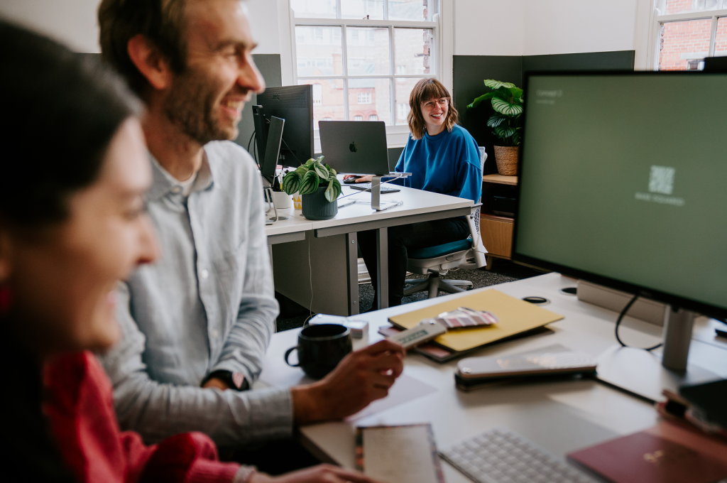 Smiling woman in a blue sweater at a desk talking with two blurred coworkers in a bright office