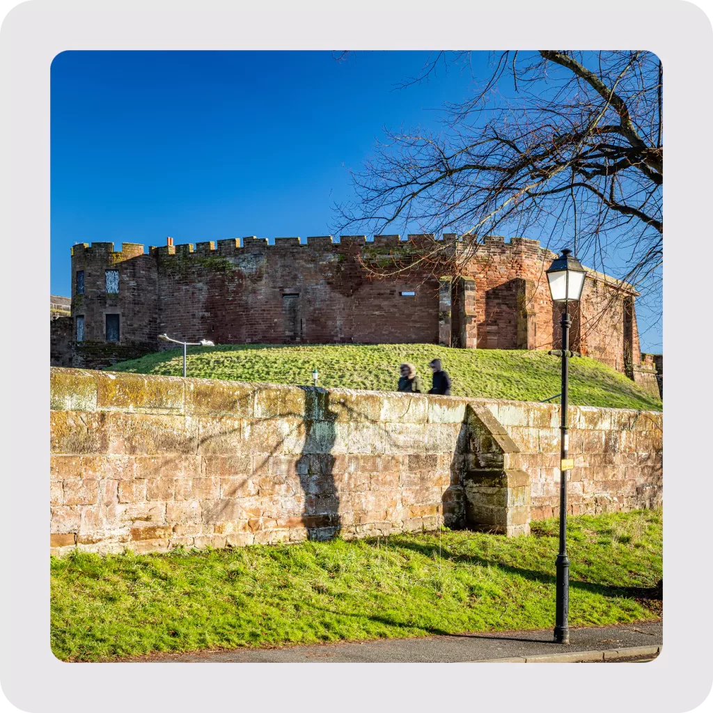 Red sandstone castle on a grassy mound above a stone retaining wall with a lamppost and two blurred walkers