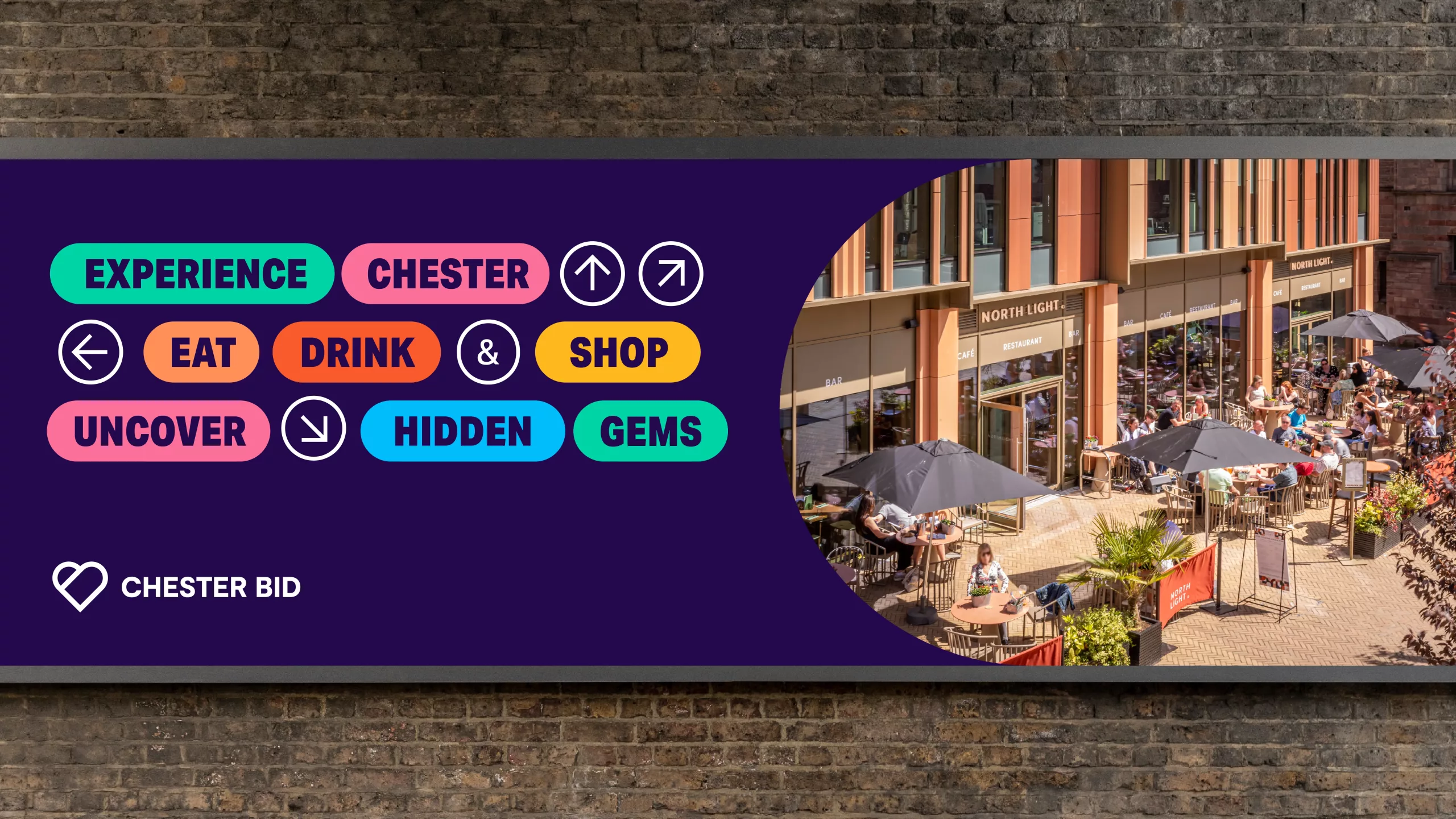 Billboard on brick wall with colourful Experience Chester text and pill-shaped Eat, Drink & Shop badges beside a circular photo of a sunny outdoor café
