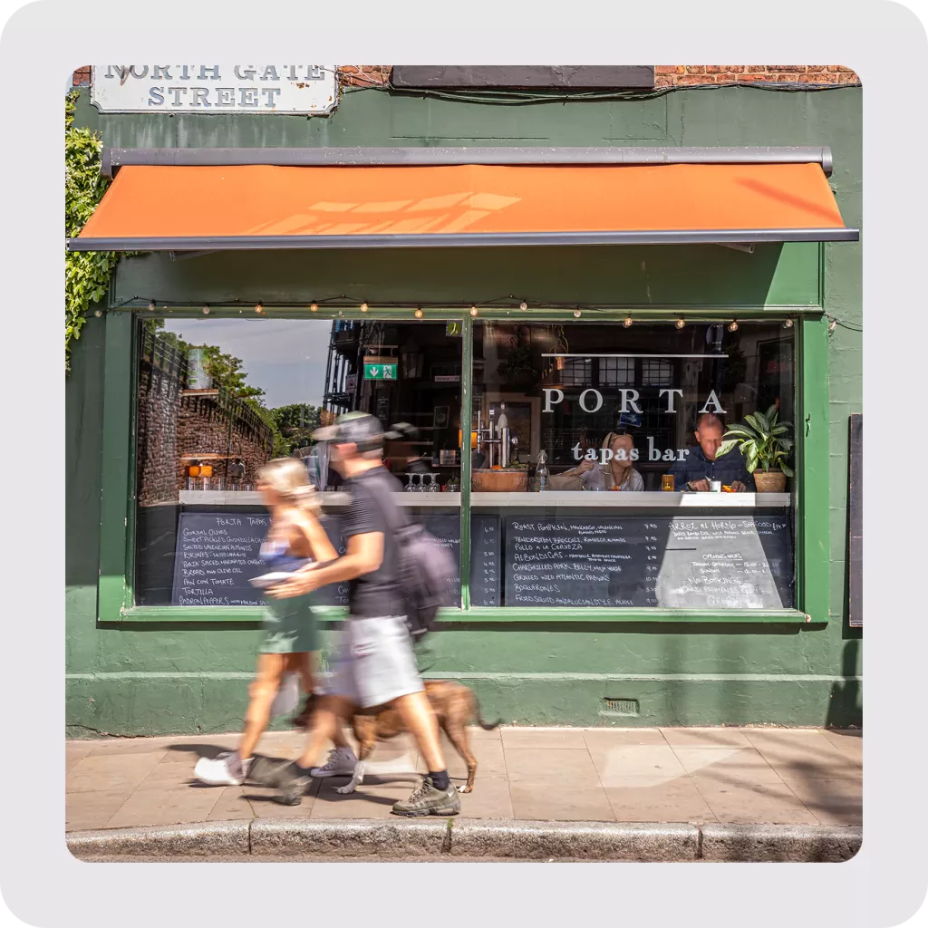 Green tapas bar Porta with orange awning, chalkboard menus in the window and blurred pedestrians walking a dog past.