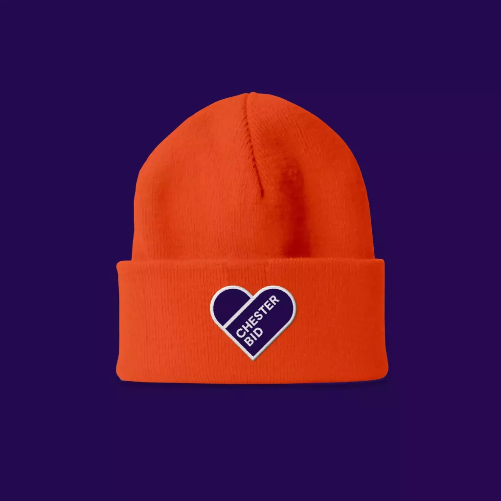 Bright orange knit beanie with folded cuff and heart-shaped purple patch reading CHESTER BID on a dark purple background.