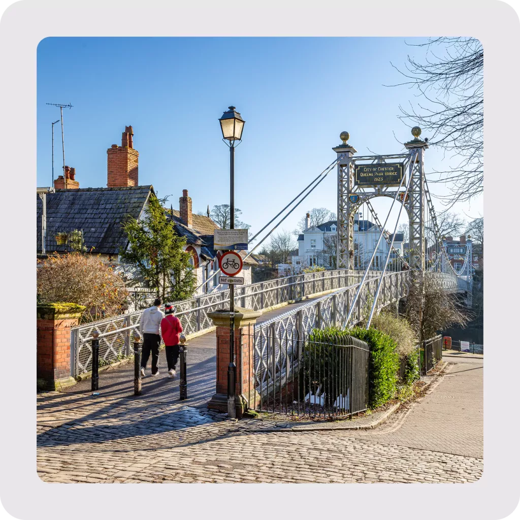 Ornate iron suspension footbridge crossing a river, lamp post at the entrance and two people walking from a cobbled street