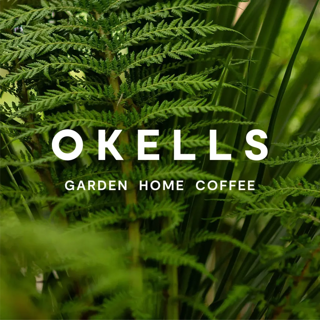 Lush green fern fronds with bold white text OKELLS and smaller text GARDEN HOME COFFEE overlaid