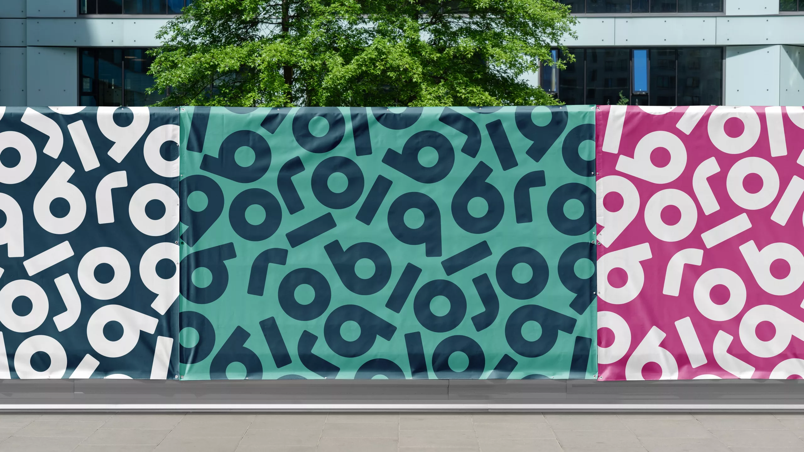 Three colourful banners (navy, teal, magenta) with repeating circular-and-bar motifs, set against a tree and office facade.