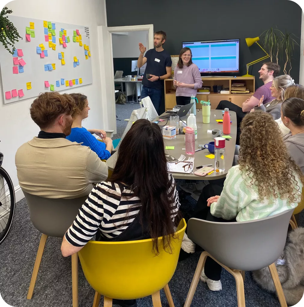 Office team meeting with people around a table listening to two presenters, sticky-note board and screen visible.