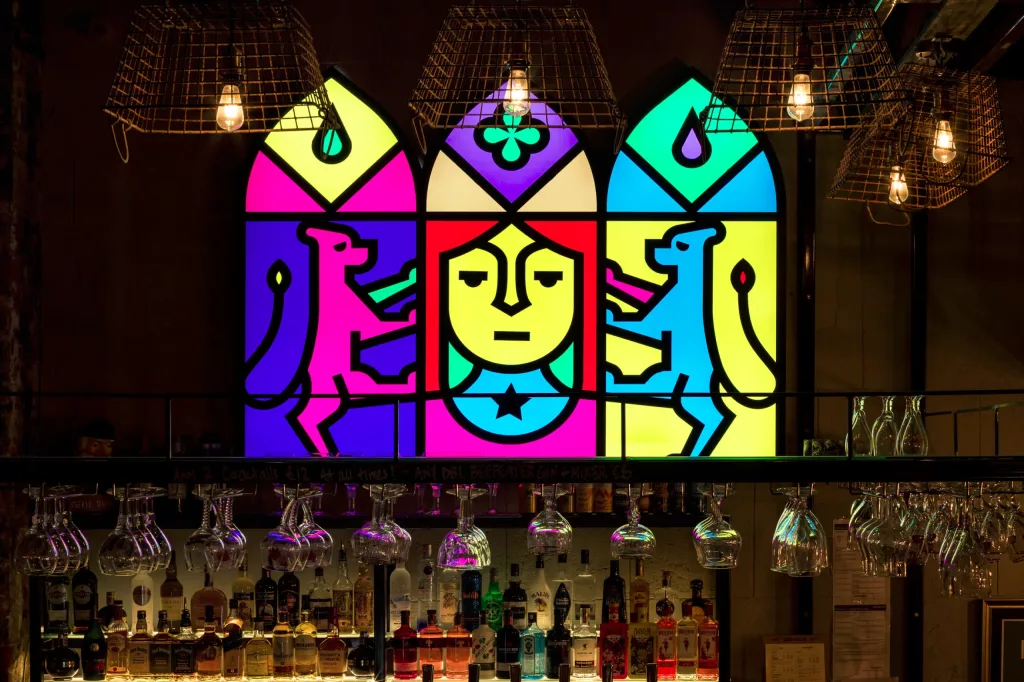 Neon stained-glass panel above a bar with a central stylised face flanked by pink and blue animals and hanging lights.