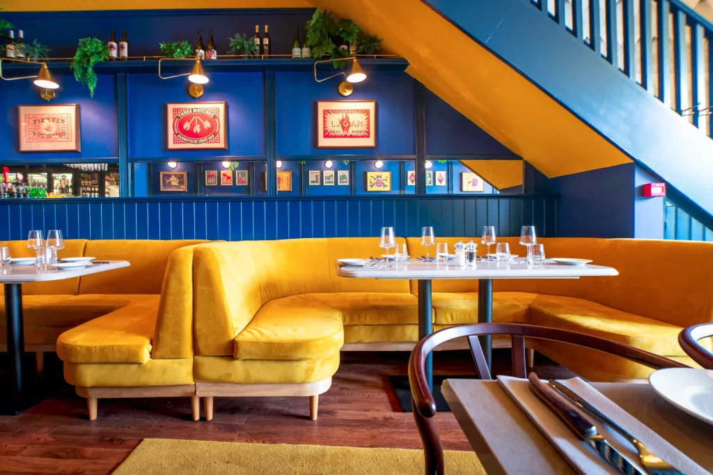 Mustard-yellow velvet banquette seating and white set tables against deep blue panelled wall with framed art and staircase.