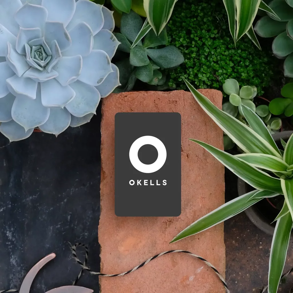Dark card with white circular logo and OKELLS text perched on a red brick amid succulents and variegated houseplants.