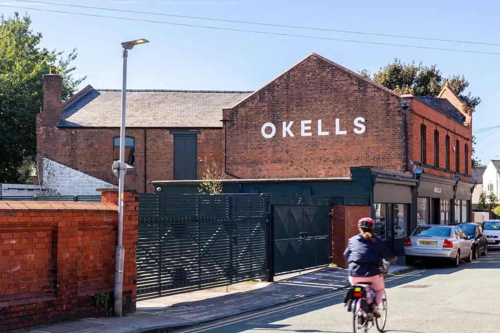 Red-brick building with large white OKELLS sign on the gable, dark metal gate and a cyclist riding past on the street.