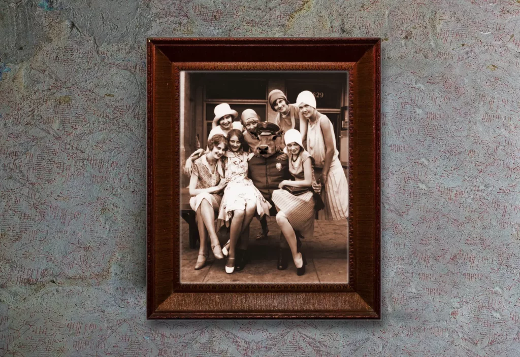 Framed sepia photograph of 1920s flapper women gathered around a police-uniformed figure wearing a bull mask.