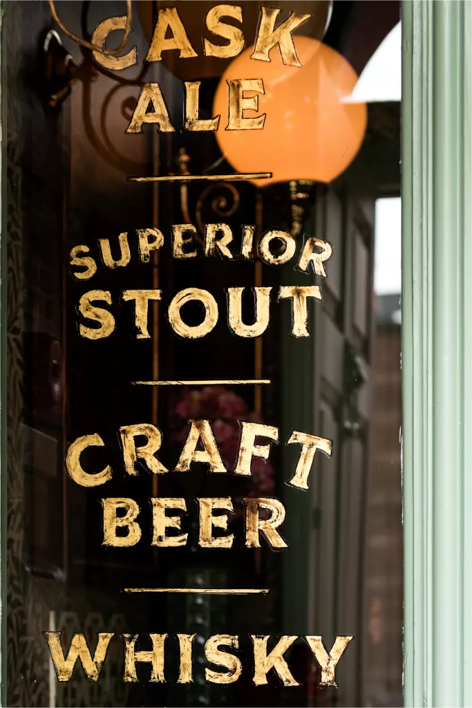 Gold-painted pub window lettering reading Cask Ale, Superior Stout, Craft Beer and Whisky, with a warm lamp reflected behind it.
