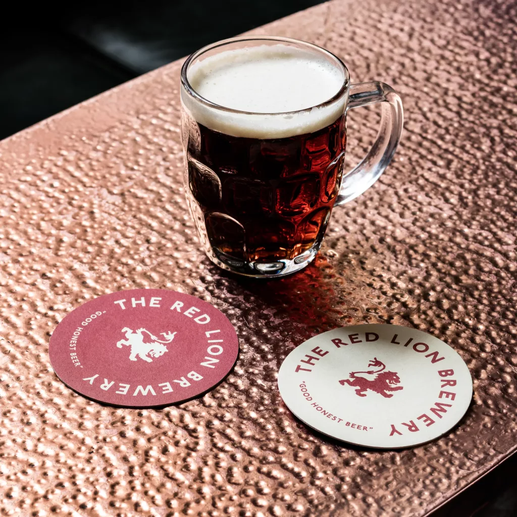 Glass tankard of dark amber beer with a creamy head on a hammered copper table beside two Red Lion Brewery coasters.