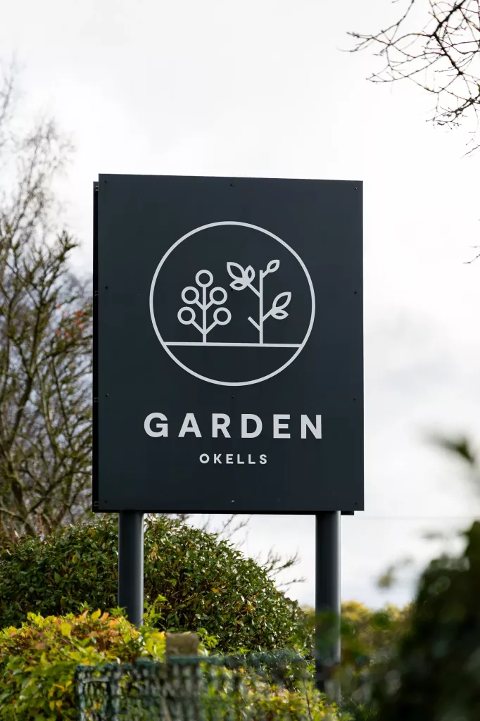 Dark rectangular sign reading GARDEN OKELLS with white circular plant logo mounted on posts above garden shrubs.