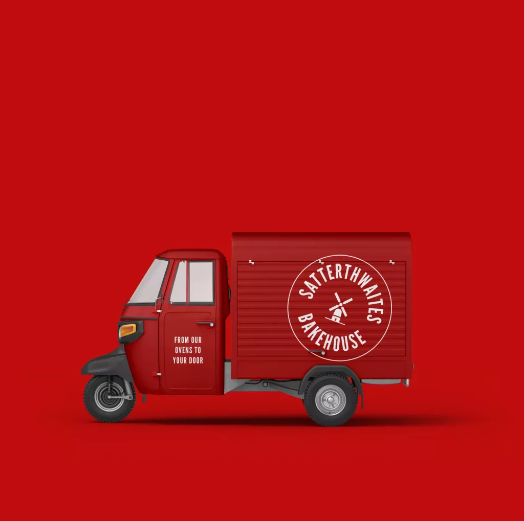 Red three-wheeled delivery van with white Satterthwaites Bakehouse windmill logo and the slogan From our ovens to your door.