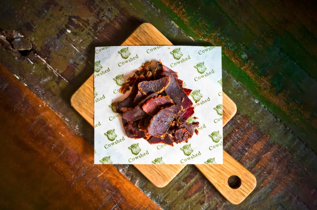 Sliced dark cured beef biltong piled on Cowshed branded paper atop a small wooden cutting board.