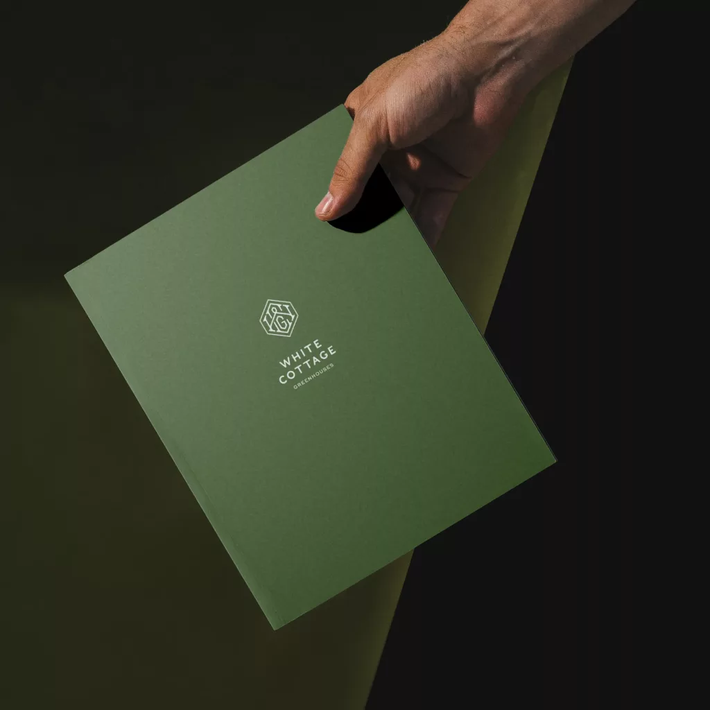 Hand holding a dark green folder with a white hexagon logo and the text White Cottage Greenhouses