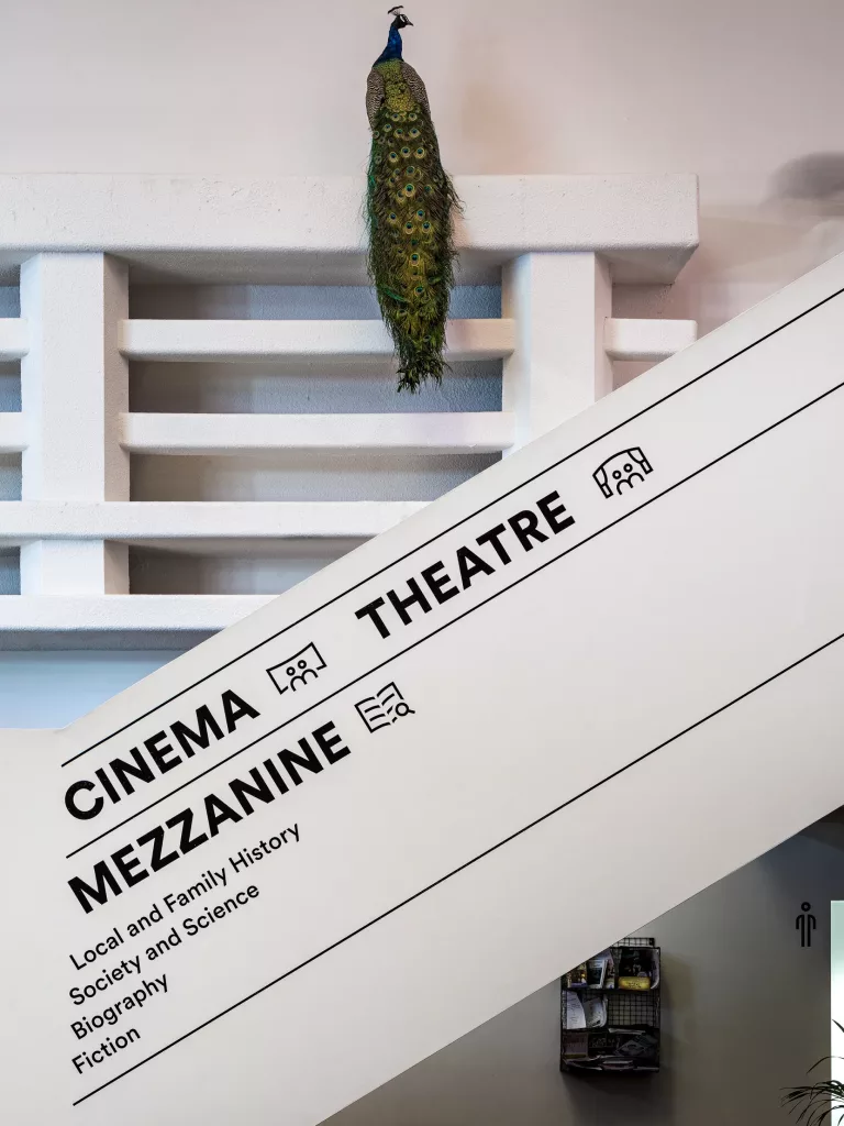 Peacock perched on a white interior ledge with long green tail draping above a diagonal wall sign reading CINEMA THEATRE MEZZANINE