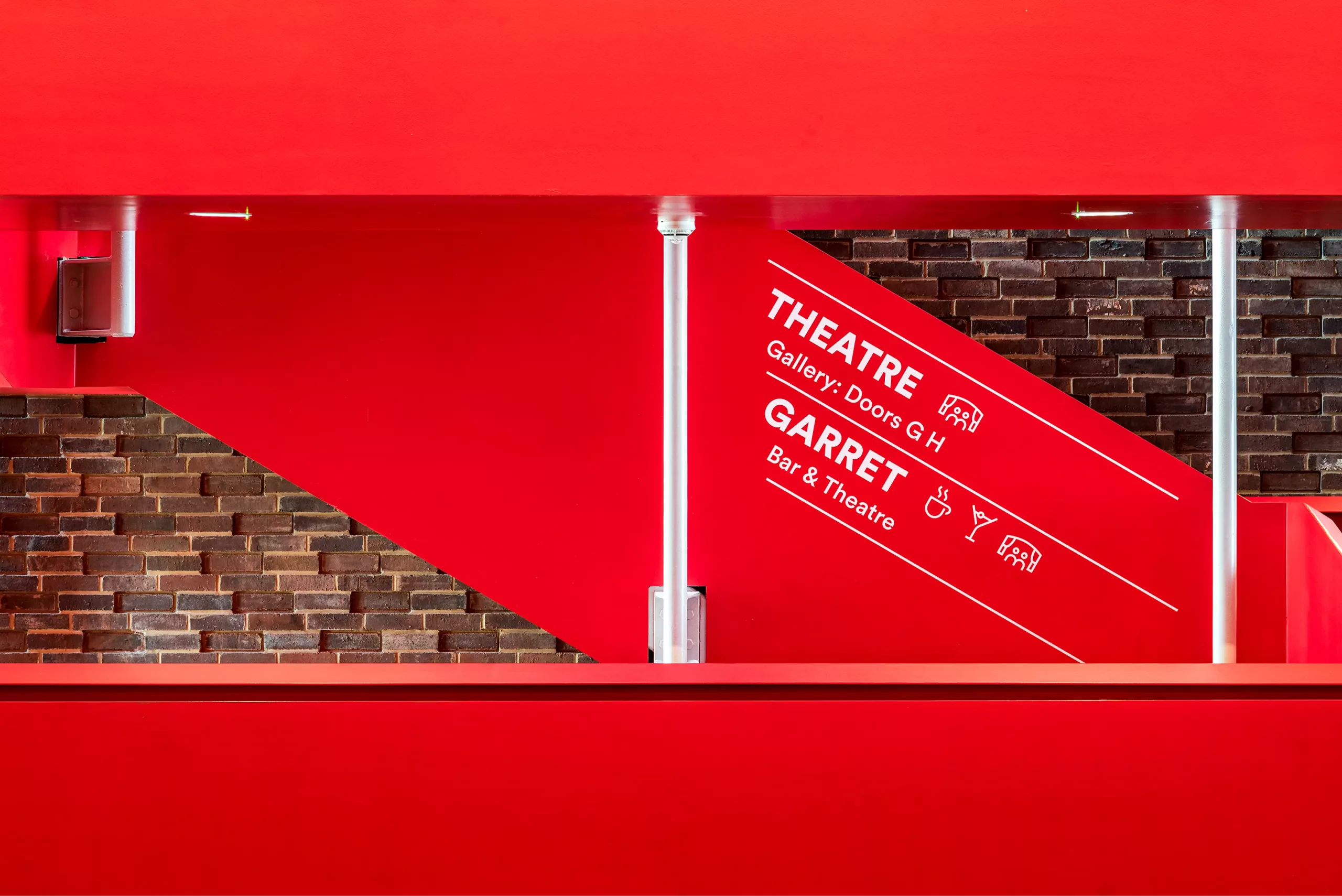 Bold red interior stairwell with diagonal white Theatre signage, exposed dark brick wall and vertical silver posts.