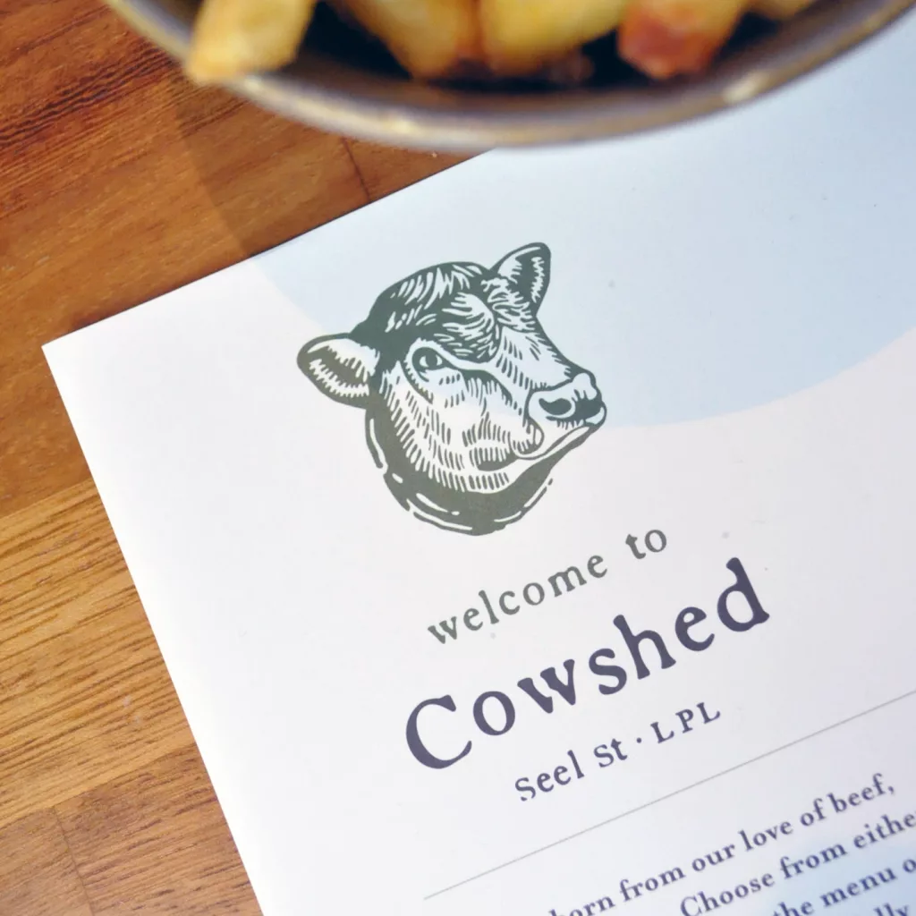 Restaurant menu on a wooden table showing an engraved cow head logo, the words welcome to Cowshed, and a bowl of fries.