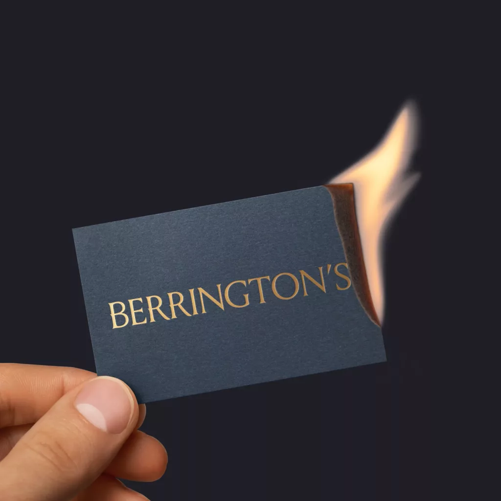 A hand holds a dark blue business card embossed with gold BERRINGTON'S, its right corner burning with a small flame