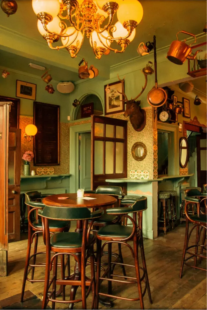 Cosy vintage pub interior: wooden high table with green leather bar stools beneath a brass chandelier and mounted deer head.