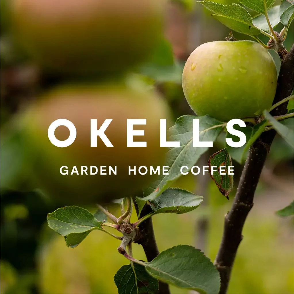 Green apple with water droplets on a tree branch, blurred apples behind, overlaid text OKELLS Garden Home Coffee