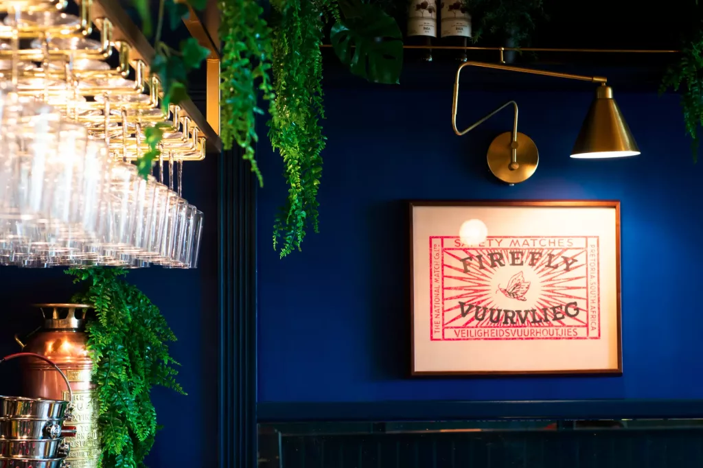 Deep blue bar wall with framed vintage matchbox print lit by brass swing lamp, hanging wine glasses and trailing green plants