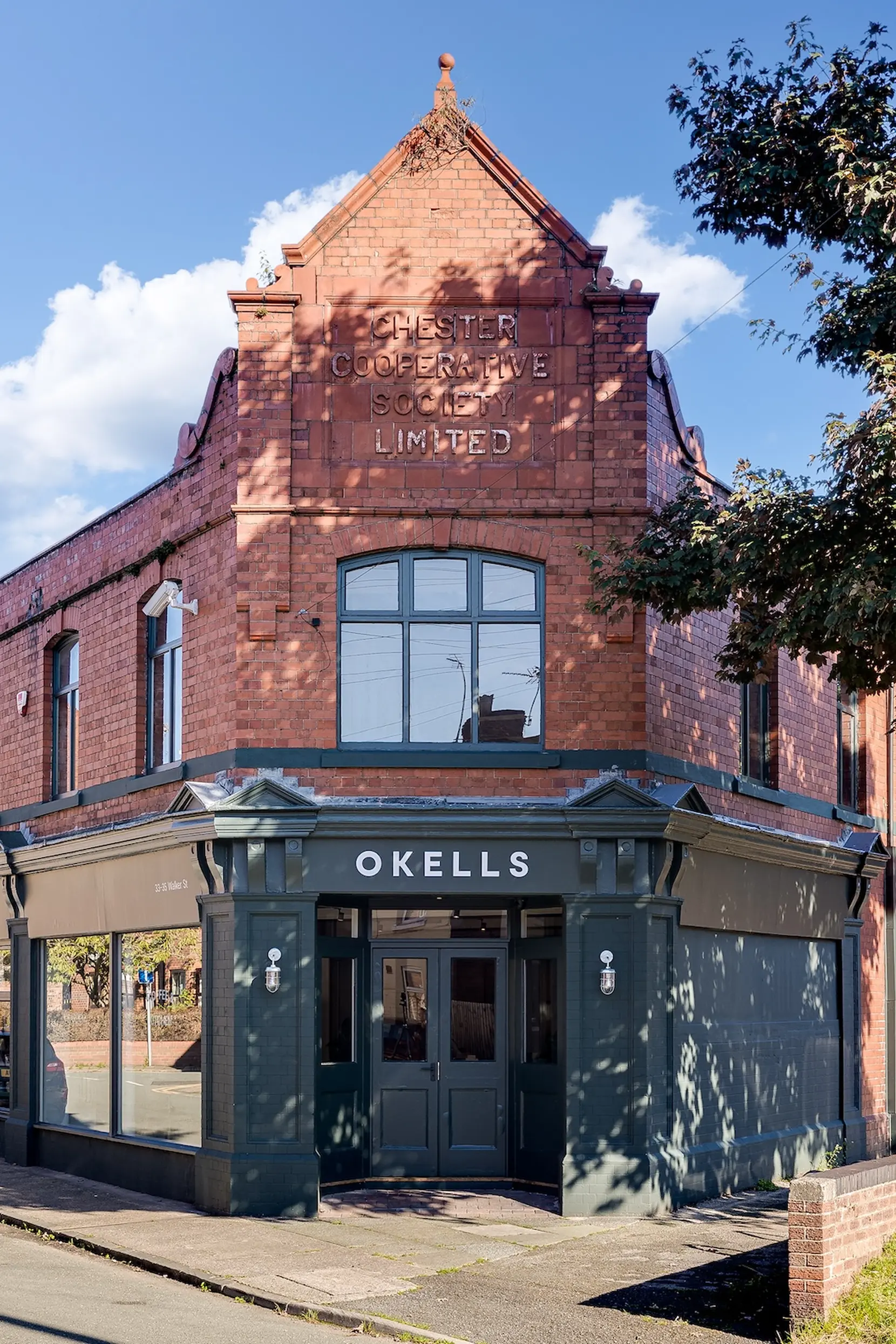 Red brick corner building with carved Chester Cooperative inscription and dark green ground-floor frontage reading OKELLS