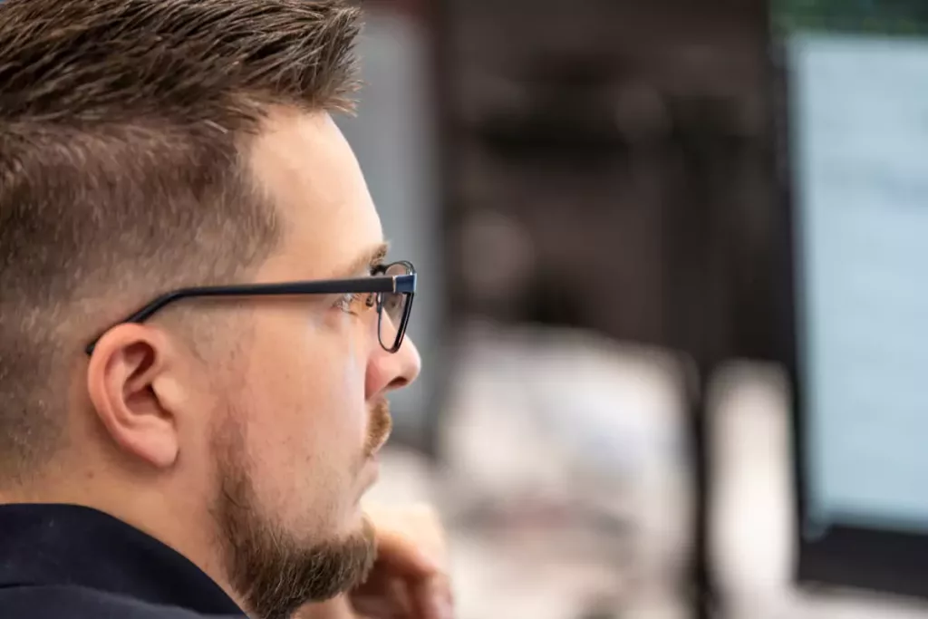 Man with short hair and glasses in side profile, resting his chin and looking at a blurred computer screen.