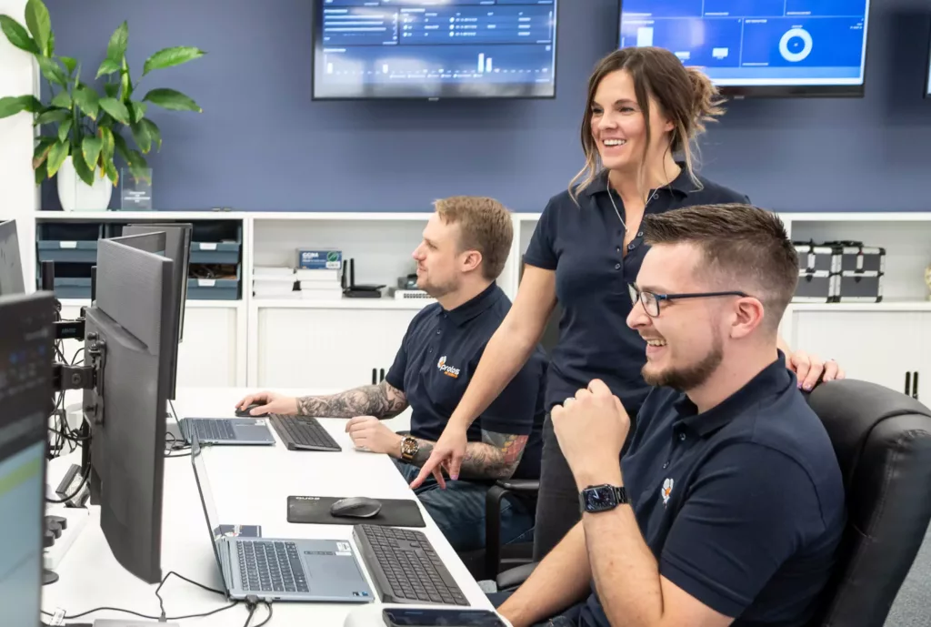 Three colleagues in navy polos smiling and working at monitors in an office, one woman standing and pointing at a screen.