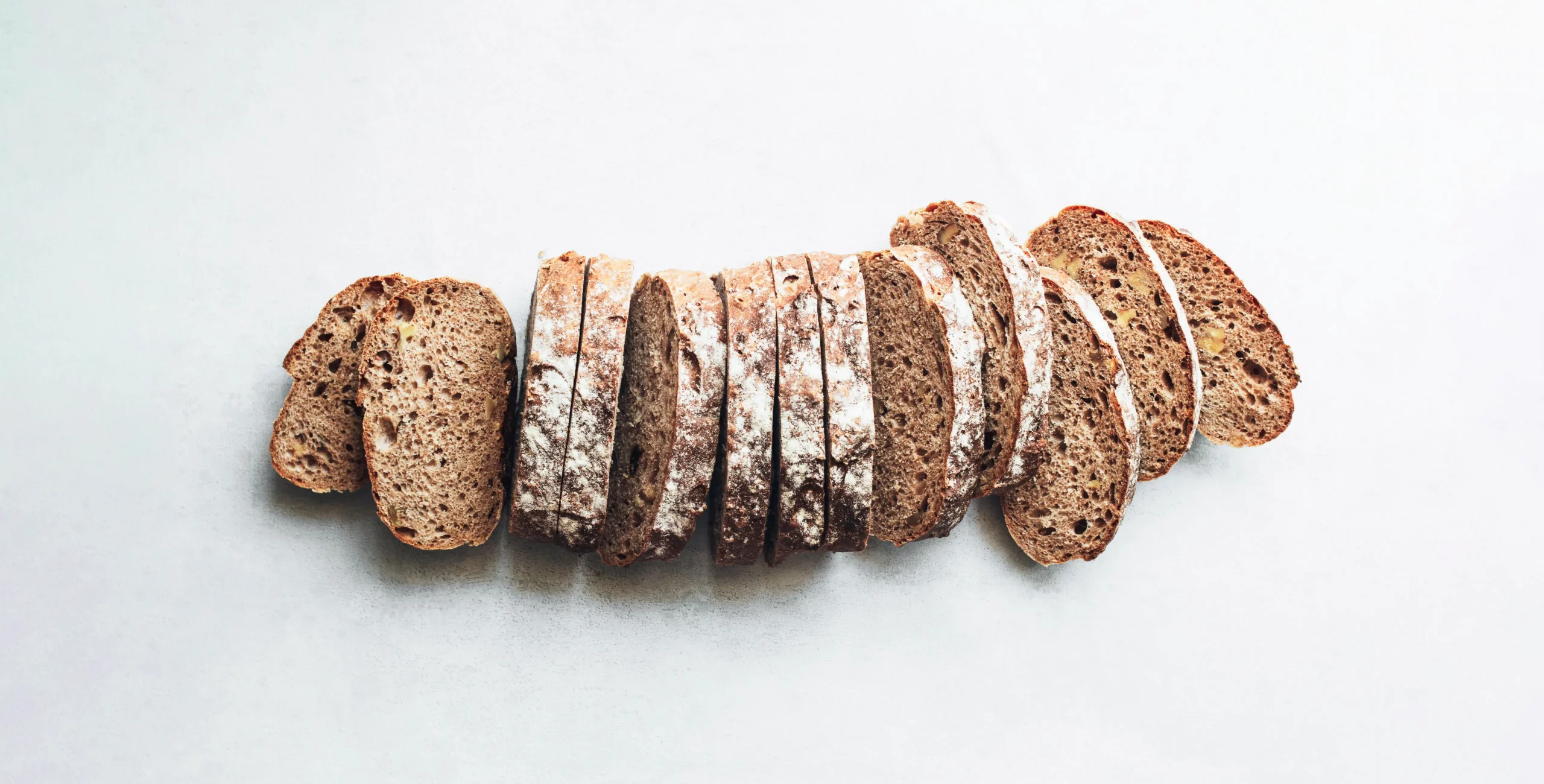 Sliced rustic wholegrain loaf dusted with flour, showing open crumb, arranged horizontally on a pale background.