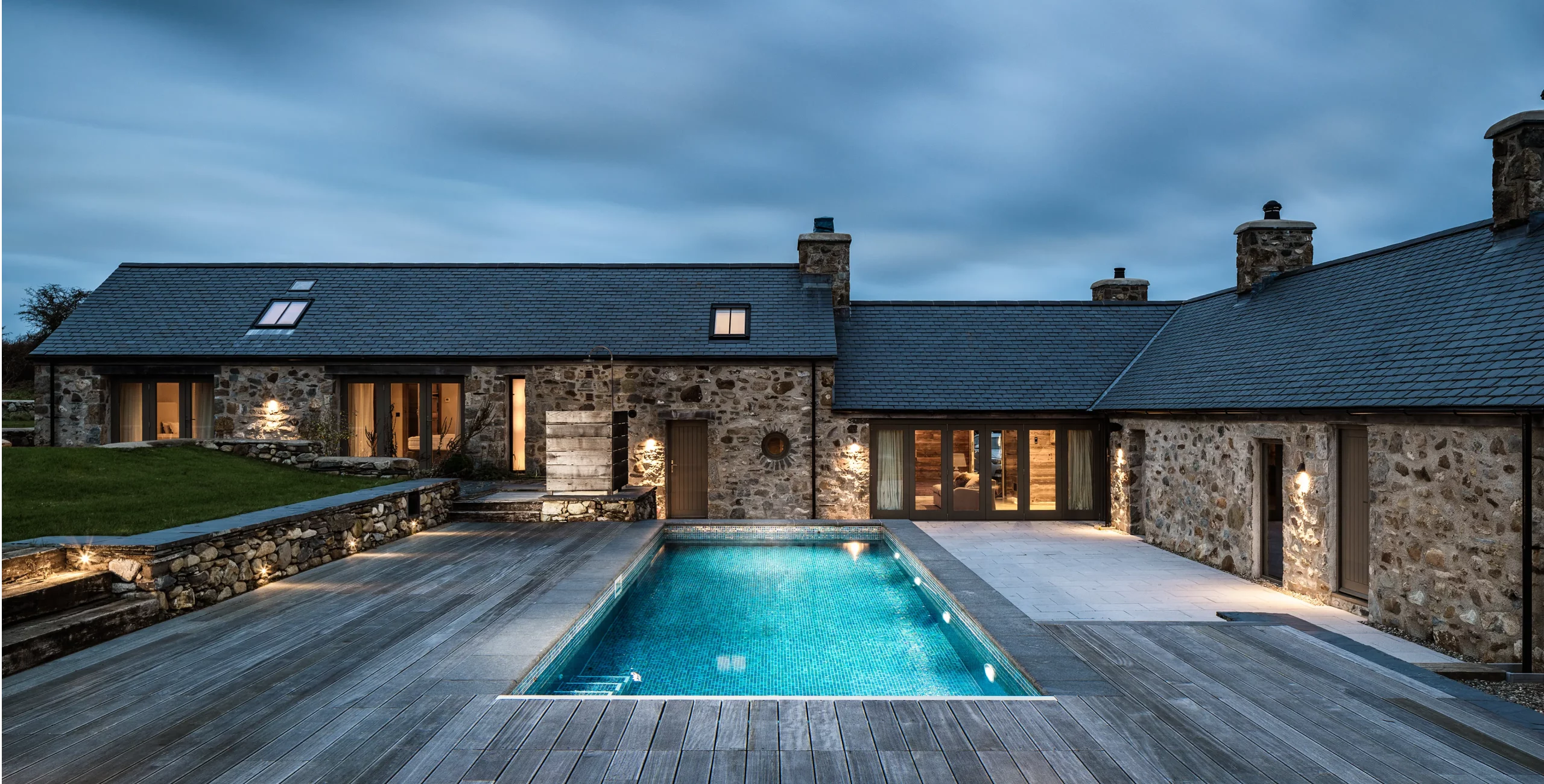 Stone country house at dusk with illuminated rectangular swimming pool and wooden decking in the foreground.