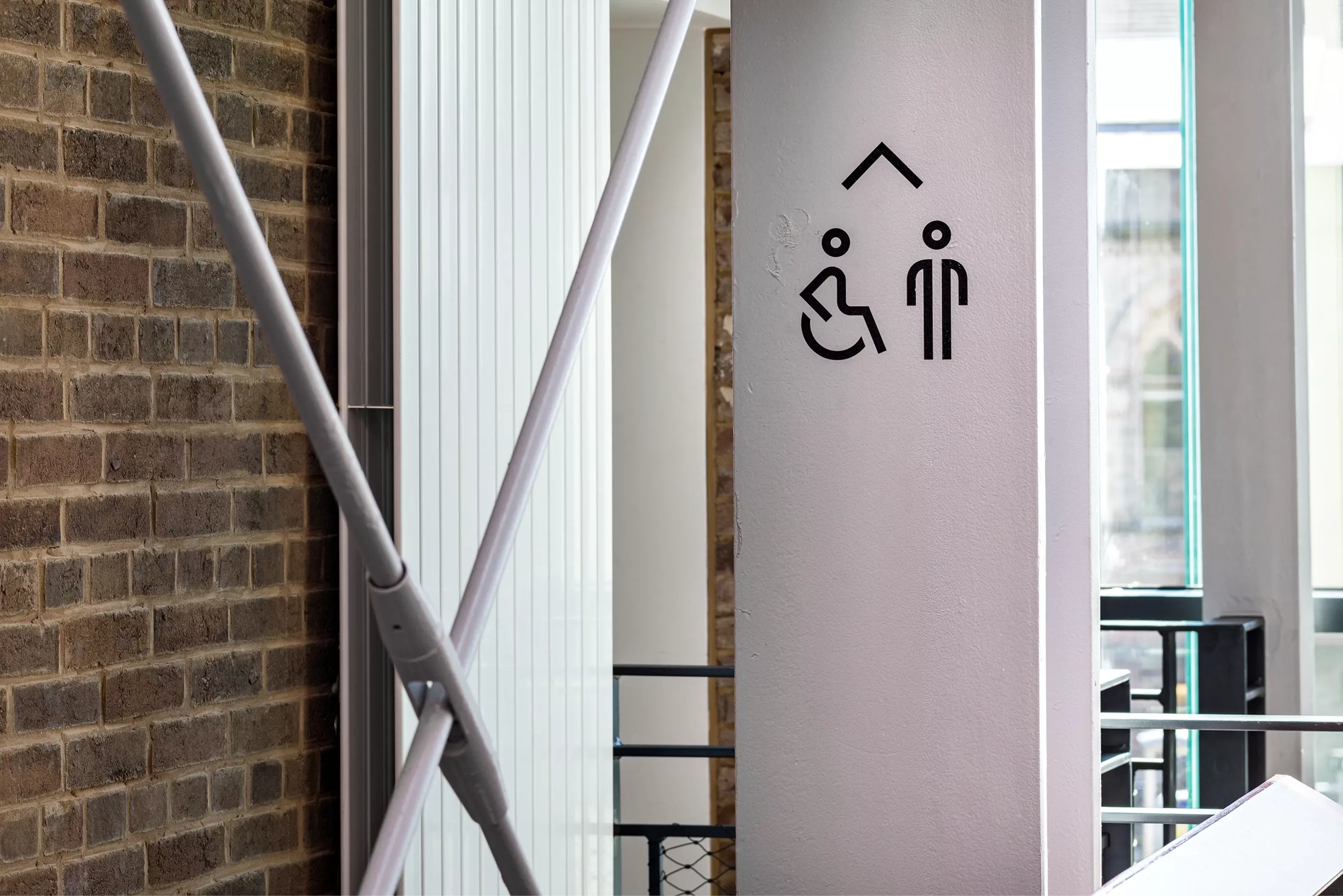 Black pictogram of a wheelchair user and standing person with an upward arrow on a pale column inside a building beside a brick wall and metal supports.
