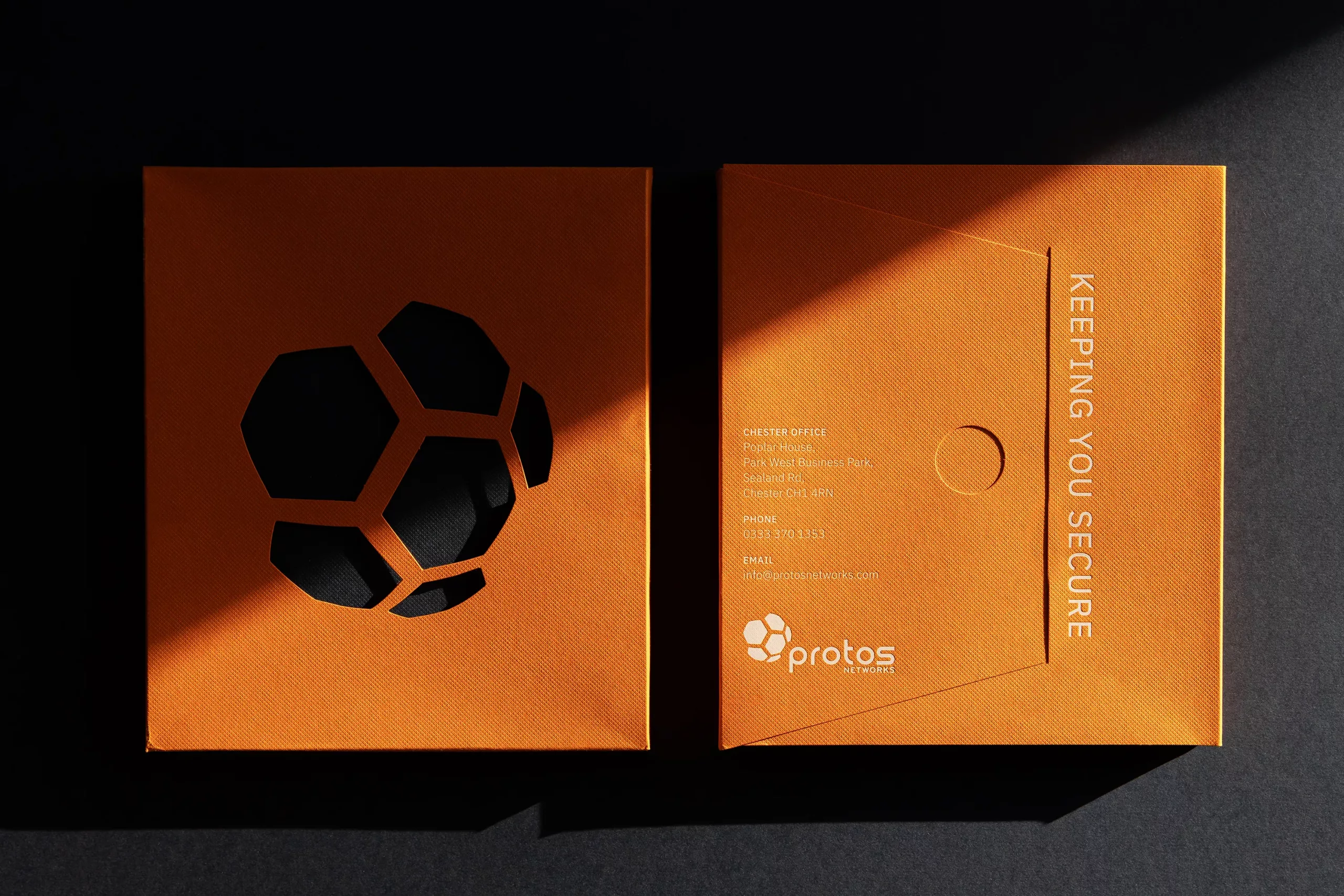 Two orange boxes on a dark surface; left has a black geometric cutout, right bears protos logo and KEEPING YOU SECURE