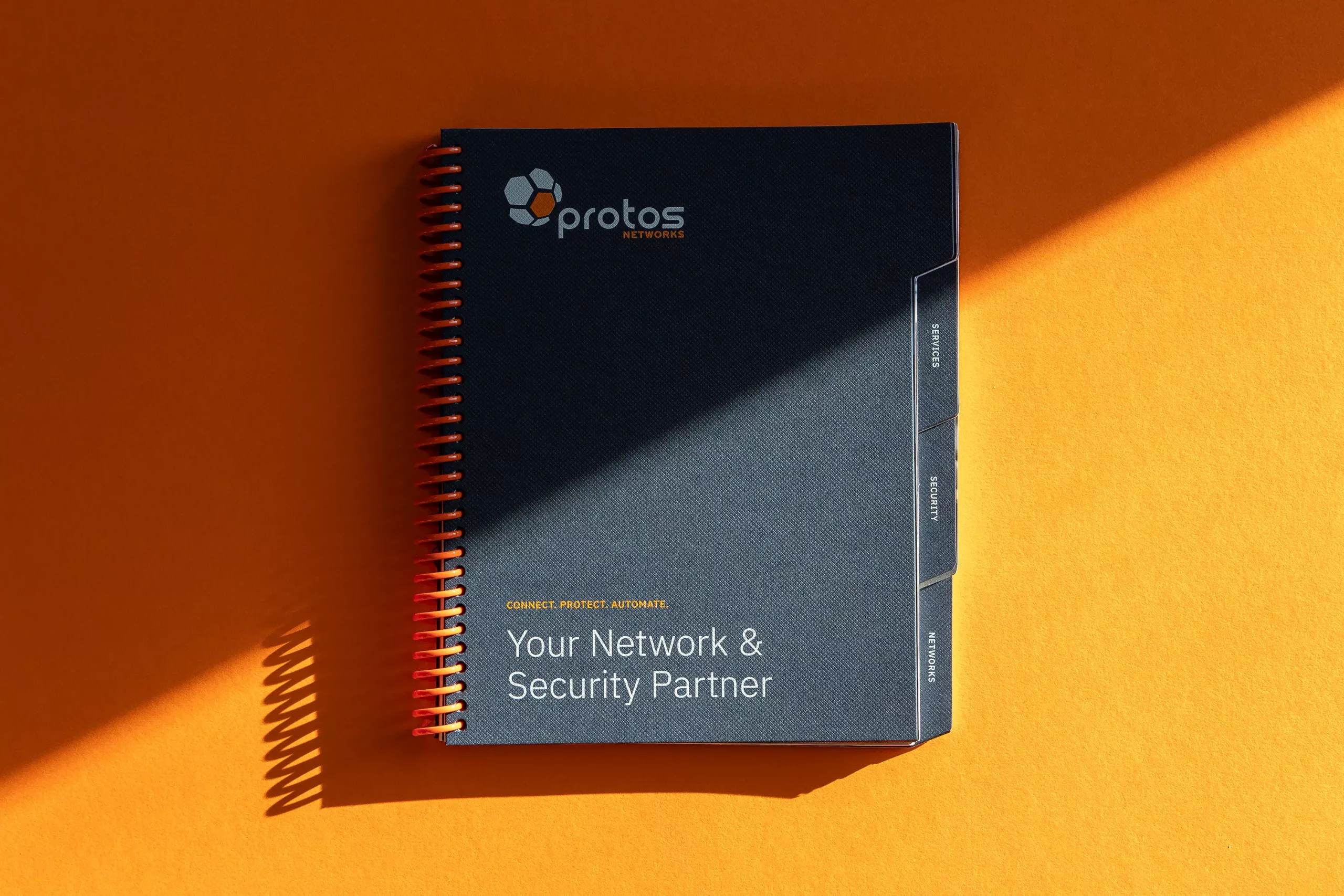 Navy spiral notebook with Protos Networks logo and tabs titled Your Network & Security Partner on orange background.