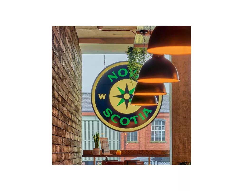 Round hanging 'Nova Scotia' compass sign seen through a café window with three glowing pendant lights and a brick interior.
