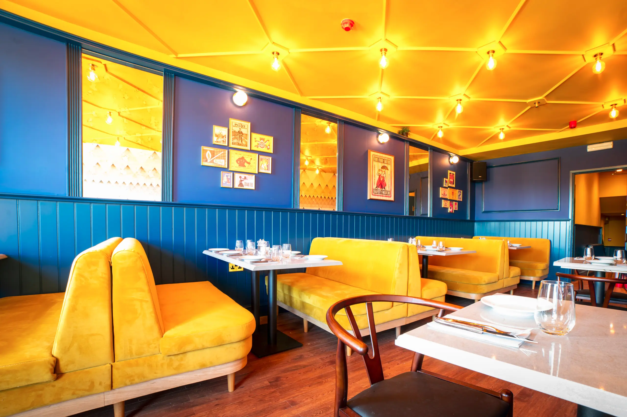 Vibrant restaurant interior with mustard-yellow banquettes, deep blue paneled walls, a warm yellow ceiling with exposed bulbs and neatly set white tables