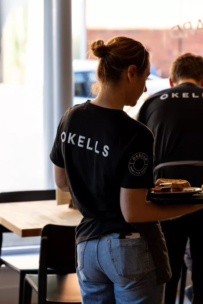Rear view of a café server with hair in a bun wearing an OKELLS black t-shirt and jeans, carrying a tray with a sandwich.