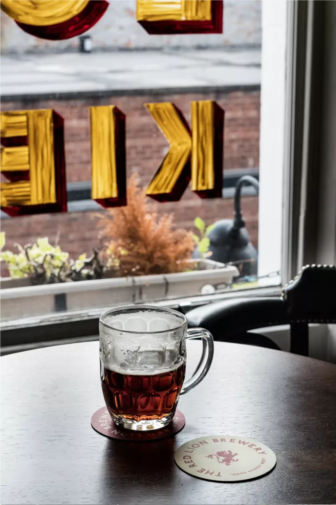 Half-full dimpled beer mug on a wooden table on a coaster reading Red Lion Brewery, window with gold reversed letters behind.