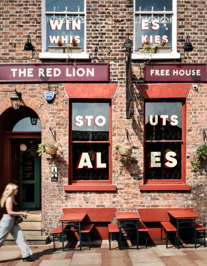 Brick pub Red Lion with red-trim windows spelling STOUTS/ ALES, hanging baskets and outdoor benches; a blurred woman passes.