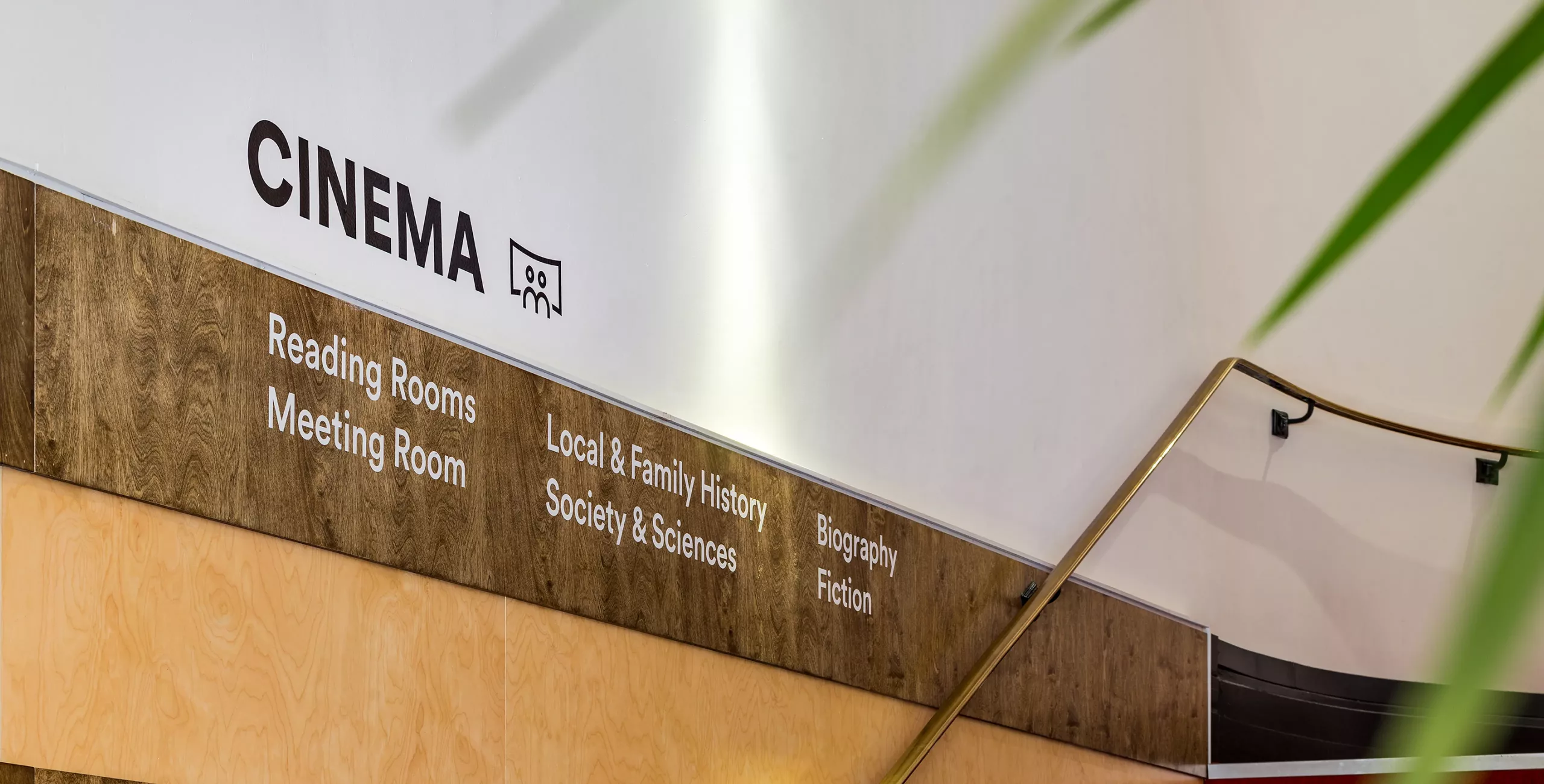 Curved staircase with brass handrail beside a wood-paneled wall signed CINEMA, listing sections such as Reading Rooms, Biography and Fiction.