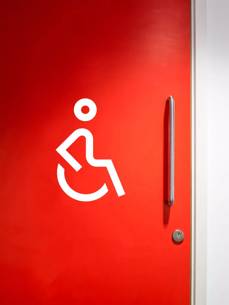 Red door with white wheelchair-accessible symbol, long stainless-steel vertical handle and round lock.