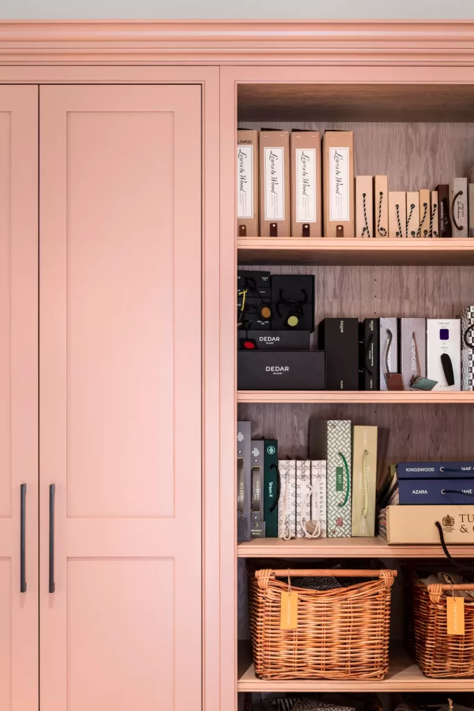 Built-in pastel pink cabinet with black handles and open shelves holding binders, fabric samples and wicker baskets.