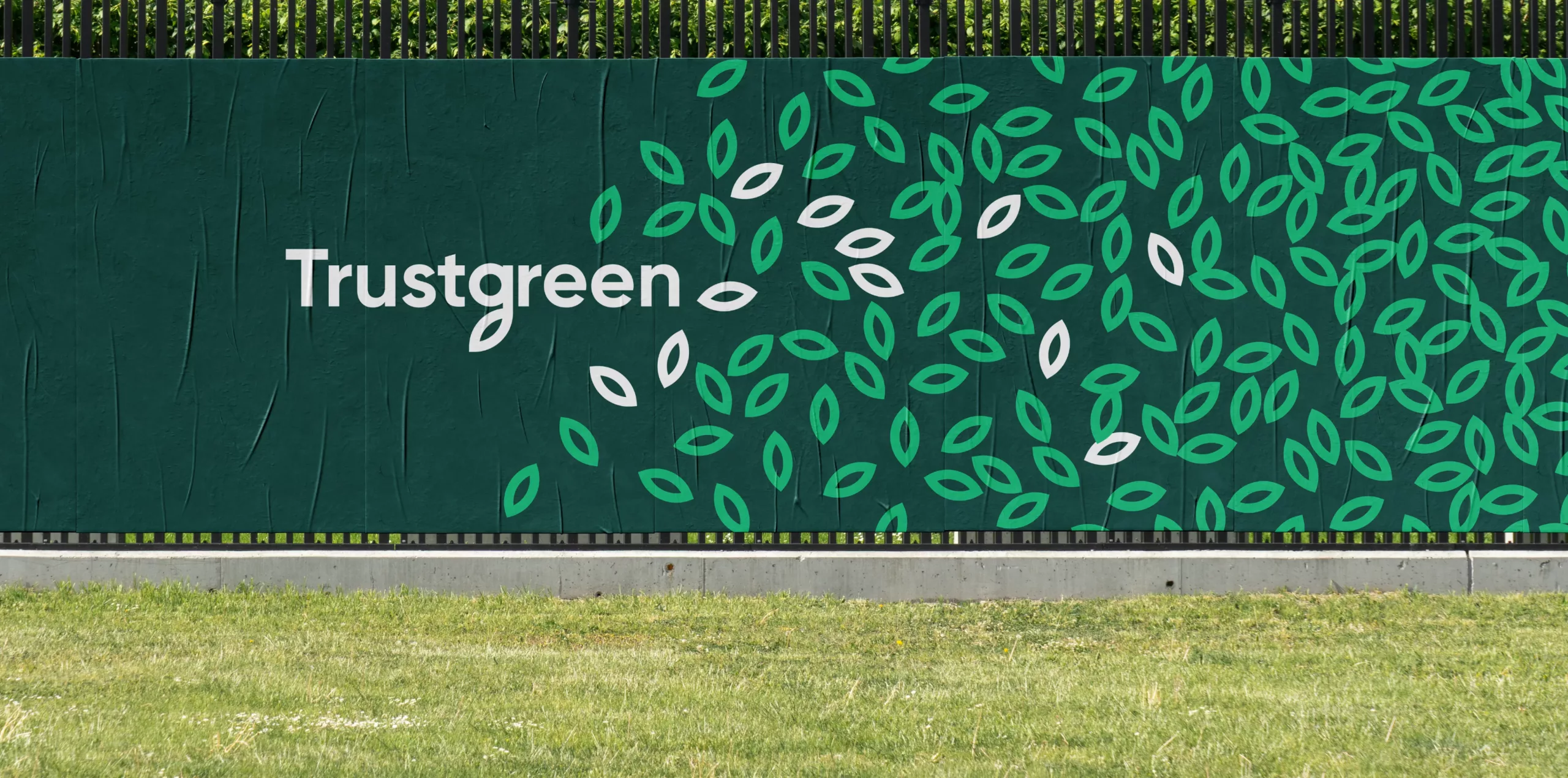 Dark green hoarding reading Trustgreen with scattered green and white leaf motifs above a grassy verge.
