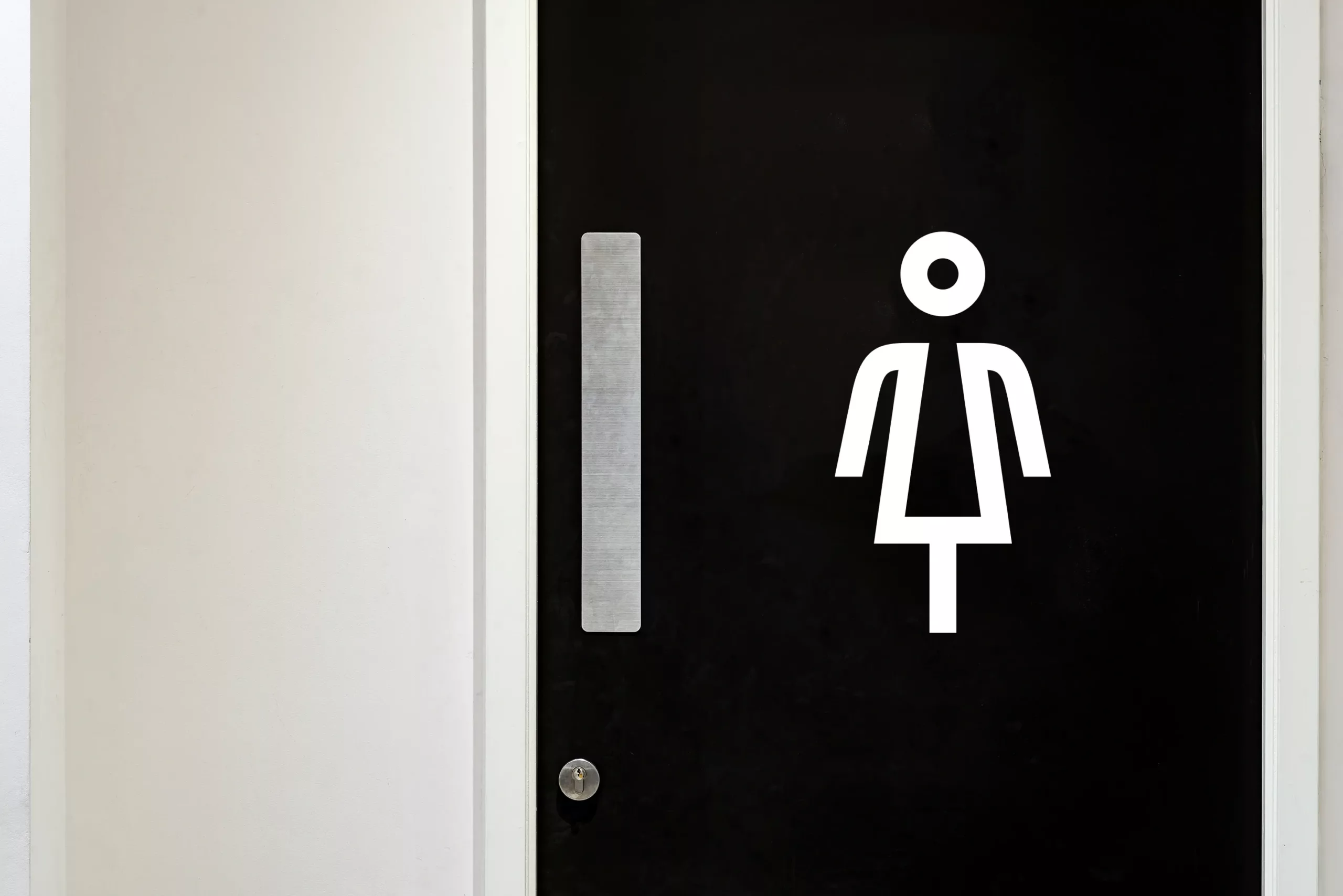Black door with white stylised women's restroom symbol, vertical brushed-metal pull and small round lock set in a white wall.
