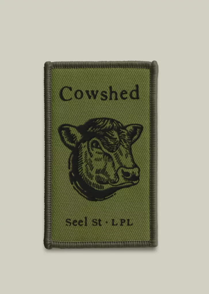 Rectangular olive-green embroidered patch with black cow head illustration and the text Cowshed above and Seel St · LPL below.
