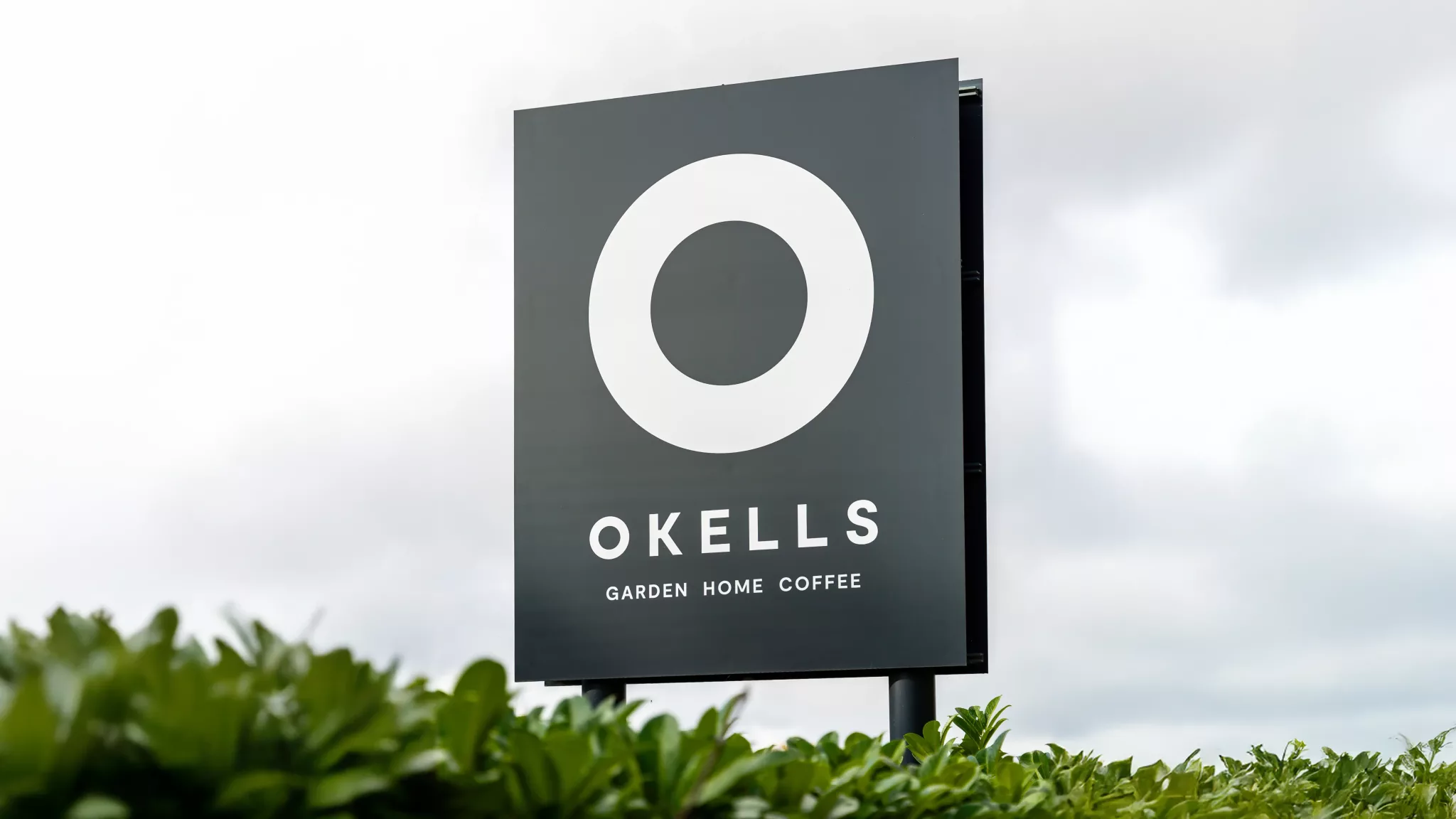 Tall black sign featuring a large white O above the word OKELLS and Garden Home Coffee, hedge in the foreground.