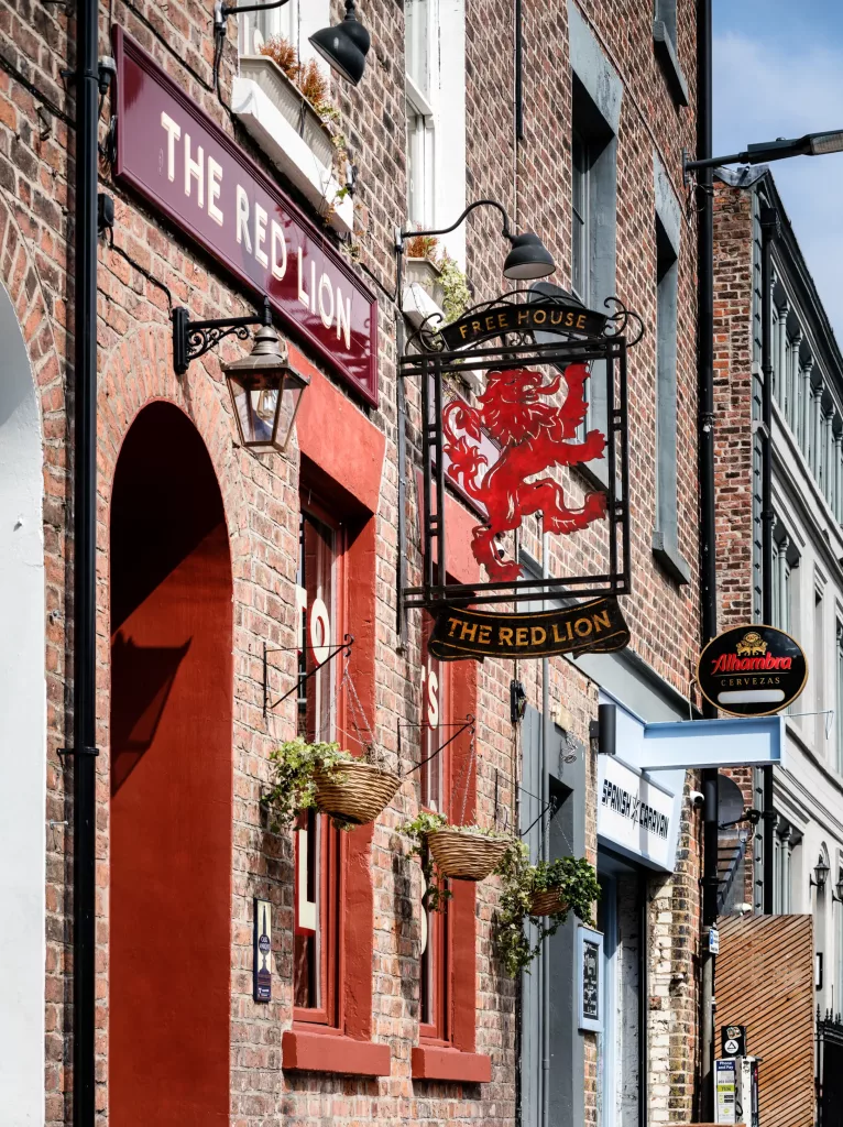 Brick pub façade with maroon sign The Red Lion, wrought-iron hanging sign showing a red lion and three plant baskets.