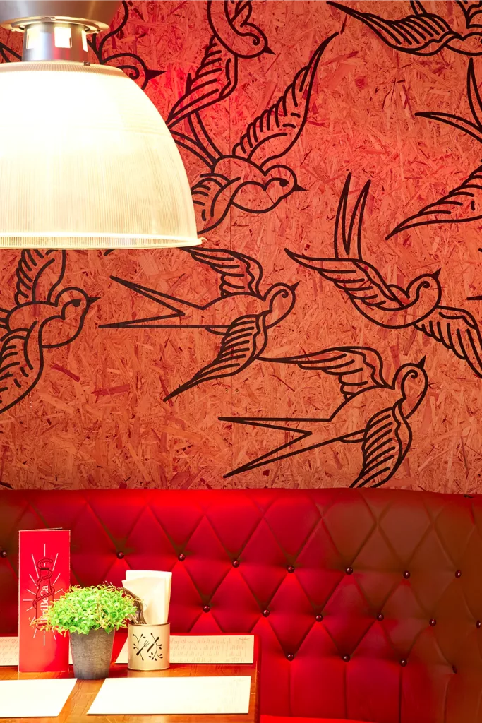 Red tufted booth beneath a wooden wall with black swallow drawings, lit by a pendant lamp over a table with a plant and menu.