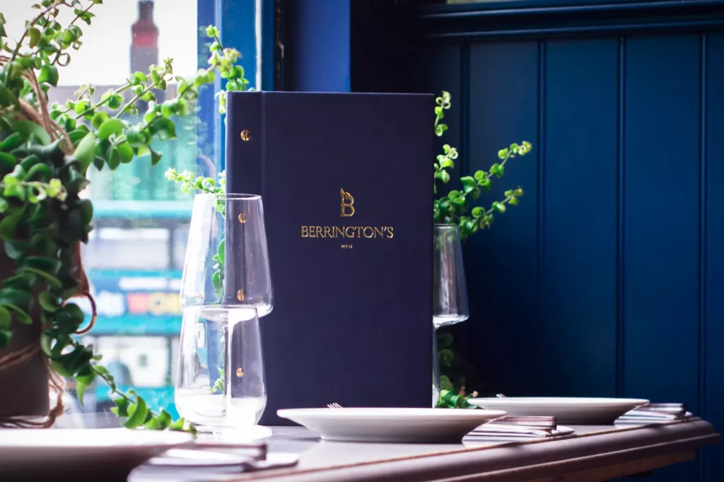 Dark blue menu with gold BERRINGTON'S text on a table between wine glasses, plates and trailing green plants by a window.